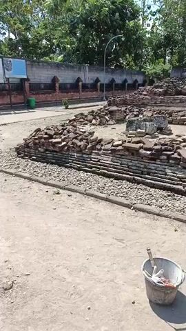 The video pans across the ruins of an ancient structure made of brick. In the foreground, a bucket with tools and possibly construction debris sits on the dusty ground. Behind the ruins, a line of trees and a wall can be seen. In the center of the ruins, a statue of Buddha is housed under a small wooden shelter with a red roof. The sky is blue with some clouds, and the overall setting appears to be outdoors in a sunny, tropical climate.