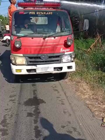 A red fire truck with "POS KUTARBLANC" visible on its side is parked on a dirt road. A firefighter in an orange uniform is seated in the driver's seat. Another firefighter in a blue uniform stands on top of the truck, operating a water cannon that sprays a powerful stream of water. The water is directed towards the right side of the frame, where lush green vegetation and palm trees are visible. A red and white striped utility pole stands in the foreground. The scene appears to be outdoors under a clear sky, possibly in a rural or semi-rural area.