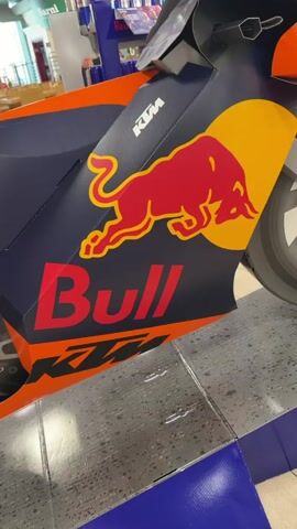 The video shows a close-up, side-view of a display for a KTM motorcycle. The motorcycle display is predominantly dark blue with orange accents. Prominently featured is the Red Bull logo, a charging red bull superimposed on a yellow circle, and the word "Bull" in large red lettering with an orange outline. The KTM logo is also visible in white. The motorcycle display appears to be made of cardboard or a similar material, with a front wheel visible. The display is on a patterned floor, with blue and purple borders at the bottom. In the background, there are tables and chairs, suggesting a public space like a cafe or food court. Some people are visible in the background, seated at tables. There are also shelves with Red Bull products in the background. The overall impression is a promotional display in a commercial setting.