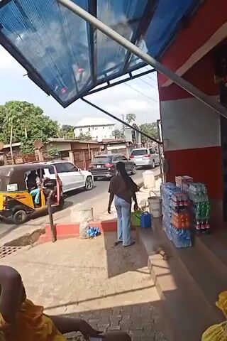 The video shows a street scene from under the awning of a small store. The street is busy with cars and a three-wheeled vehicle known as a "Keke Napep" or auto-rickshaw. A woman in jeans and a brown top is standing on the steps of the store, carrying a blue container. Another person, partially visible, is seated and wearing a yellow top. Stacks of bottled water and soda are on display on the store steps. The environment appears to be a sunny day in a residential area.