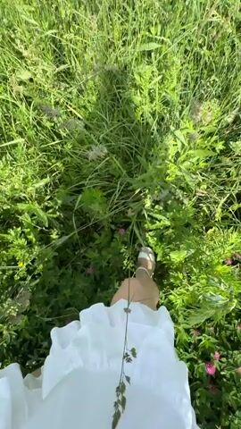 The video shows a first-person perspective of someone walking through tall green grass and vegetation. In the background, there are rolling hills with scattered trees and a bright blue sky with scattered clouds. A dirt road is visible in the distance with a couple of cars driving on it. The person appears to be wearing light-colored shoes or boots. The camera is shaky, indicating movement. The environment is rural and appears to be a sunny day.