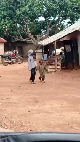The video shows a dusty, outdoor marketplace. Several goats are present, some near a maroon car with its trunk open. A red motorcycle is parked nearby. In the background, there are buildings with corrugated metal roofs and several people are visible, some standing and others seated. A line of motorcycles is parked under a yellow umbrella. A woman in a purple headscarf carries a bag on her head. A man in a green outfit is speaking into a mobile phone. The overall impression is of a rural or semi-urban setting with everyday commerce taking place.