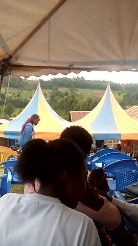 The video shows a group of people sitting under a tent. They are sitting on plastic chairs, and some of them are looking at their phones. Outside the tent, there is a view of green hills and trees. The tent is supported by wooden poles. There is another smaller tent in the background.