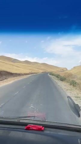 The video captures a view from inside a vehicle, looking forward through the windshield. The road ahead is paved and appears to be a two-lane highway winding through a hilly, arid landscape. The hills on either side of the road are covered in dry, yellowish grass. The sky is blue with scattered white clouds. There is a side mirror visible on the right side of the frame, attached to the vehicle's exterior. On the dashboard, in front of the windshield, a small red object is placed. The camera is stationary, suggesting it is mounted within the vehicle. The overall scene depicts travel on a highway in a dry, possibly mountainous region.
