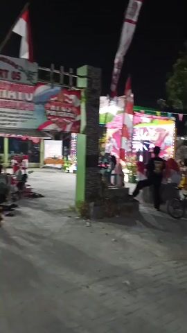 The video shows an outdoor gathering at night. The scene is illuminated by artificial lights and decorated with Indonesian flags and colorful banners. Several people are present, some sitting on chairs and others standing. In the foreground, a person with long dark hair is smoking. In the background, there appears to be a stage or performance area with more decorations and people. The ground is paved with bricks and concrete. The overall atmosphere suggests a celebratory or community event.