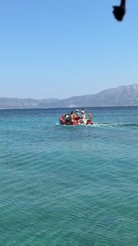A small red inflatable boat with several people on board is moving through clear blue water. The boat is creating a wake behind it. In the background, there are distant hills and mountains under a clear blue sky. The water appears shallow in the foreground, revealing the sandy bottom.