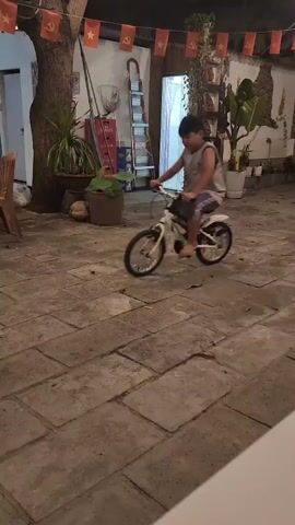 A young boy wearing a grey sleeveless shirt and blue shorts rides a white bicycle on a paved courtyard. The boy pedals the bike as it moves forward. In the background, a woman sits at a table. The courtyard is decorated with flags.