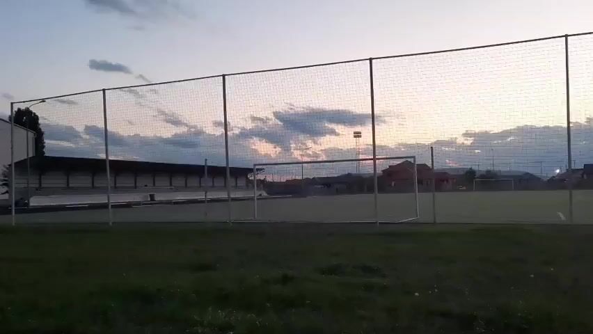 This media file captures an outdoor scene at dusk, likely in Gudermes, Russia. The primary subjects are a sports field with soccer goals and a running track. The setting appears to be a sports complex or school grounds, with buildings visible in the background. The sky displays a gradient of blues and grays, with scattered clouds, indicating the transition from day to night. A few lights are visible on tall poles in the distance, suggesting artificial illumination for the sports facilities. The overall mood is serene and quiet, characteristic of early evening. There is no visible text within the media file.