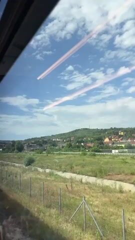 The video is filmed from inside a moving vehicle, likely a train, looking out a window. The window has reflections of internal lights, appearing as pinkish-white streaks across the top half of the frame. Outside, a rural landscape unfolds under a blue sky with scattered white clouds. In the foreground, a patch of dry grass and a wire fence with thin posts are visible. Beyond the fence, a larger area of green grass or crops stretches towards a village. The village consists of numerous houses with red-tiled roofs, nestled on the side of a gently sloping hill. Trees are interspersed among the houses, and the entire scene is bathed in daylight. The camera perspective suggests the vehicle is moving from left to right relative to the landscape.