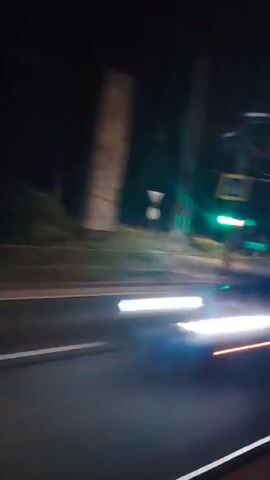 A blurry night-time video shows a view from a moving vehicle. The video captures a road with white striped markings and a pedestrian crossing. Streetlights illuminate the scene. Trees and utility poles are visible in the background. The vehicle appears to be moving along a highway or a well-lit road.
