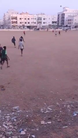 In a wide shot, several people are playing soccer on a dirt field in an urban environment. Buildings are in the background. The sky is overcast and a strip of garbage litters the lower edge of the frame. The soccer players are casually dressed and moving around the field. One man is walking toward the viewer, carrying a bundle of cloth. The overall impression is of a recreational activity taking place in a somewhat unkempt setting.