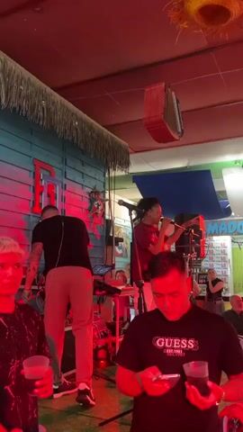 The video captures an indoor live music performance, likely at a bar or casual venue in Fuengirola, Spain, during the evening. The main subject is a male singer with dark, curly hair, wearing a maroon t-shirt, who is energetically performing on a small stage. He holds a microphone and gestures with his free hand, engaging with an unseen audience.

Behind the singer, another male musician is visible from the back, operating a keyboard or DJ controller setup, suggesting a live band or accompaniment. The stage area is illuminated by dynamic red and green lights, which cast strong hues on the performers and immediate surroundings. The backdrop of the stage features a light blue wooden wall, possibly designed to look rustic, with a thatched overhang. A decorative red object, resembling an accordion or a patterned speaker cover, hangs from the ceiling.

In the foreground, two male audience members are visible. On the right, a man wearing a black t-shirt with "GUESS EST. 1981" printed on it holds a clear glass with a dark beverage and looks down at his phone. To his left, another man with light-colored hair also holds a drink. In the background, beyond the stage, several other people are present, and signs displaying "LA TEVA" and multiple instances of "5.0" are visible, possibly indicating prices or a brand name. The overall scene depicts a lively and relaxed atmosphere for a musical event.