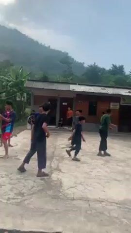 Here's a forensic description of the video:

The video appears to be shot in a rural or semi-rural area, possibly a village. In the initial scene, a group of approximately 10 young males are visible, seemingly playing a game or engaging in some form of physical activity on a concrete platform in front of a single-story building. The building appears to be a simple structure, possibly a school or community center, with a corrugated metal roof. Lush green vegetation is visible in the background, including hills and trees. 

As the video pans, the camera reveals more of the surrounding environment. There are several small, makeshift structures or shacks made of wood and corrugated metal sheets. Two scooters are parked by the roadside. A red vending stand that seemingly offers prepared foods is also visible. The road is a dirt or gravel road. The overall impression is one of a developing or less affluent area.