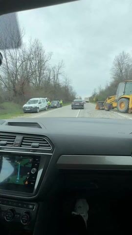 The video shows a view from inside a car driving on a road. The dashboard of the car is visible in the foreground, with a navigation screen displaying a map. Outside the car, there is a line of vehicles stopped on the road. There are three people wearing yellow vests standing near the vehicles. A construction vehicle is parked on the side of the road. The surrounding environment consists of trees and foliage. The sky is overcast.