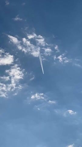 The video starts with a view of a clear blue sky dotted with fluffy white clouds. A jet plane is flying high, leaving a prominent white contrail behind it. The camera then pans down to reveal a scenic landscape. The sky is now a mix of blue and gray, with sunlight streaming through the breaks in the cloud cover. A mountain range is visible in the distance, partially shrouded in mist.