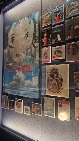 The video displays a museum exhibit featuring a collection of Studio Ghibli films. The items are encased in clear protective sleeves and mounted on a dark wall. The exhibit appears to showcase DVD or Blu-ray cases, along with posters of various films. Prominently featured are posters and cases for "Spirited Away" and "Howl's Moving Castle." The lighting in the exhibit is dim, with spotlights highlighting the displayed items. The camera pans across the wall, offering a view of numerous film titles arranged in rows and columns. Some sections of the exhibit are blurred, suggesting movement or a focus shift. The overall impression is of a curated collection dedicated to the animated works of Studio Ghibli, likely in a retail or exhibition setting.