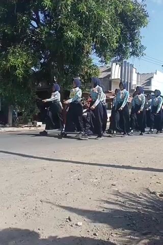 This is a video of a group of people in a line. They are all wearing similar uniforms, which include a patterned shirt, dark pants, and a blue cap. They appear to be students, possibly in a school setting. Some of them are on scooters or motorbikes. The environment appears to be outdoors, with a paved road and some trees and buildings in the background. The sun is shining, casting shadows. The people are lined up and appear to be participating in some sort of organized activity or event. Some individuals are looking forward, while others are looking down. There are multiple individuals in the frame, each seemingly engaged in their own position within the line. The video is shot from a slightly elevated angle, looking down at the scene.