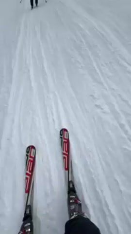 A person is skiing down a snowy slope, filmed from a first-person point of view, following another skier in front. The camera points downward to the skis, then shifts upward to reveal a mountain range in the background under a cloudy sky. The video then alternates between the skier in front and the snowy landscape, showing an overhead ski lift.