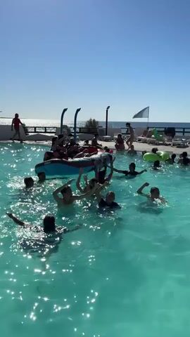 A group of children are enjoying themselves in a swimming pool on a sunny day. Some children are in the water, splashing around, while others are on an inflatable raft in the center of the pool. The children on the raft are waving and appear to be having a lot of fun. In the background, there is a blue sky, the ocean, and lounge chairs by the pool.