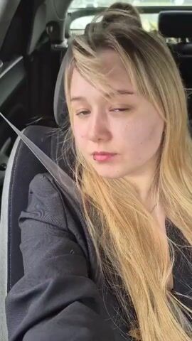 A young woman with blonde hair sits in the passenger seat of a car. She is wearing a dark jacket and a seatbelt. Her hair is long and straight, and she has a lip piercing. She turns her head to the side, then looks at the camera and smiles. The background is blurry, suggesting the car is in motion.