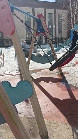 The video shows a playground in the early spring, where some snow remains on the ground. The camera is positioned at ground level, partially obscured by the wooden supports of children's swings, which feature red, blue, and natural wood colors. A child wearing a black jacket is sitting in a tire swing and lightly swaying. In the background, a person is sitting on a park bench near a metal fence. The playground has a red surface, partially covered with melting snow and water, which reflects the shadows of the swing structure. A building is visible in the background, and the sky is clear and blue.