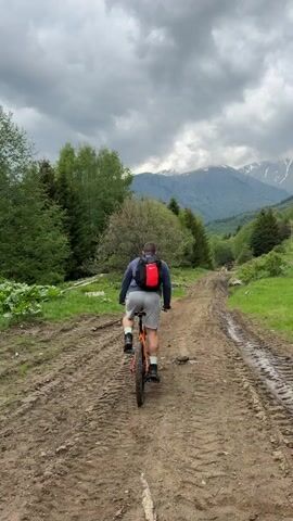 A person is riding a bicycle on a muddy dirt road. They are wearing a dark shirt, light-colored shorts, and a red backpack. The road is flanked by green grass and trees on either side. In the distance, there are mountains with patches of snow on the peaks. The sky is overcast with gray clouds. The camera follows the person on the bicycle from behind.