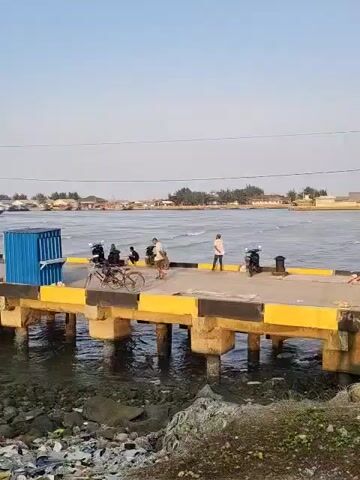 The video shows a pier jutting out into the water. Several people are on the pier, some standing and some sitting on bicycles. There are also a few motorcycles on the pier. A blue shipping container is positioned near the end of the pier. The water is choppy, and there appears to be some debris in the water near the shore. In the background, there is a coastline with buildings and trees. The sky is clear and a pale blue. The pier has yellow and black markings along its edges. The video is shot from a slightly elevated position, looking down at the pier and the water. The overall scene appears to be a working harbor or dock area.