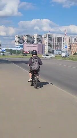A person wearing a black helmet, a dark t-shirt, and camouflage shorts is riding an electric bicycle on a paved path. The person has a gray backpack on their back. The bicycle has knobby tires, a black frame, and a red taillight. The path is next to a road with cars. In the background, there are modern buildings and trees. The sun is shining, casting shadows. Another person on a bicycle is partially visible in the foreground on the left side of the frame, and they are wearing a white t-shirt.