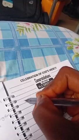 A close-up shot captures a person's hand, with a dark skin tone, actively writing with a silver pen in a spiral-bound notebook. The notebook rests on a surface covered with a blue and white checkered fabric, suggestive of an indoor setting in Ilorin, Nigeria.

On the right page of the notebook, a prominent printed insert or card displays "CELEBRATION OF GOD'S MERCY" at the top, followed by "Congratulations ON YOUR PHD CONVOCATION" within a black rectangular frame. The individual is writing on the blank lines below this message, with the partially formed words "when I a" visible under the pen.

The left page of the notebook, slightly out of focus, contains handwritten notes with phrases such as "die 200", "se of of", "resulting from", and "verse". In the blurred background, a black backpack and an orange object, possibly a container, can be seen. The scene conveys a quiet, focused moment, likely related to an academic achievement or a celebratory event like a PhD convocation.