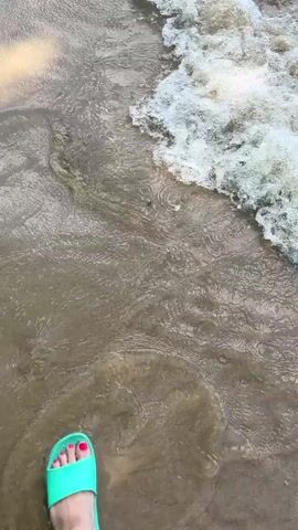 The video shows a first-person perspective of someone standing on a sandy beach with the ocean waves washing over their feet. The person is wearing a turquoise slide sandal on their left foot, and their toenails are painted pink. The water is murky brown and churns with white foam as it recedes from the shore. The lighting suggests it is daytime, with sunlight reflecting off the water.