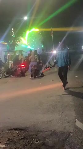 A nighttime scene unfolds on a paved road with a person walking towards the right of the frame. In the background, a crowd gathers around a stage adorned with colorful lights and speakers, suggesting an outdoor event or performance. Several motorcycles are parked on the left side of the road, some with riders seated on them. The atmosphere appears festive, with a bright light emanating from the stage area, illuminating the surrounding space and casting long shadows.