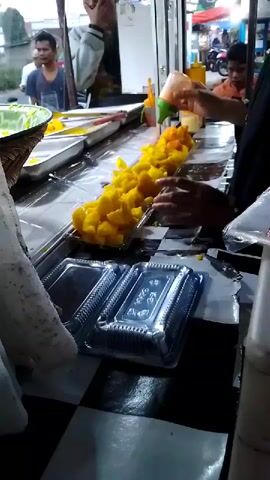 The video shows a food stall with a person in the foreground squeezing a sauce bottle over a line of yellow food items in plastic containers. There are other containers of food and bottles of sauce on the counter. In the background, two people are visible, one appearing to be a customer and the other an employee. The stall appears to be outdoors with a checkered pattern on the counter.