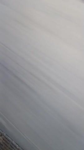 The video shows a view from a moving vehicle, possibly on a highway or a road. The road surface is gray and appears to have a white line painted on it. The motion blur suggests that the vehicle is traveling at a moderate to high speed. The lighting is consistent with daylight. There are no people or other vehicles visible in the frame. The perspective is from a low angle, looking forward and slightly down.