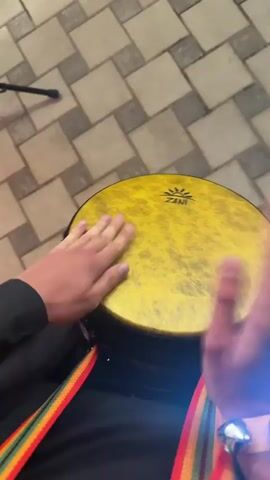 The video shows a close-up of a person's hands playing a yellow drum. The drum has the brand name "ZANI" visible on it. The person's hands are positioned over the drumhead, and the hands appear to be moving rapidly, indicating they are actively playing the instrument. A colorful strap, featuring red, yellow, and green bands, is visible hanging from the drum. The person is also wearing a yellow bracelet with small bells attached to it, which jingle as the hands move. The background appears to be an indoor setting with a tiled floor. The lighting is somewhat dim, with a bright light source visible in the upper left corner of the frame.