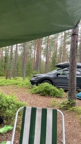 The video shows a campsite in a forest. In the foreground, a striped camping chair is visible. A green tarp is extended over the campsite, providing shelter. To the right of the chair, a dark colored car with a roof box is parked. Next to the car, an inflatable tent with blue accents and the brand name "Quechua" is set up. The tent is partially zipped open. The background consists of a dense pine forest with tall trees and lush green undergrowth. The ground is covered with pine needles and dirt. There appears to be a small clearing with a fire pit and some scattered camping equipment in the distance. The lighting suggests an overcast or cloudy day.
