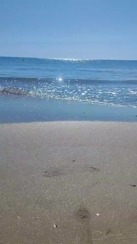 The video captures a close-up, low-angle view of a sandy beach with gentle waves lapping at the shore under a clear blue sky. The sun glints off the water's surface, creating a shimmering effect. In the foreground, a person's bare legs and feet are visible as they walk along the wet sand, leaving footprints behind. The individual is wearing dark blue swim trunks with white lettering. The camera focuses on the movement of their feet and legs against the backdrop of the ocean and sky.