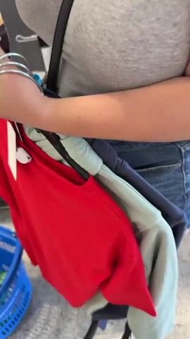 A person's hands are visible, holding a package of Nutella biscuits and a red t-shirt on a hanger. A blue shopping basket is in the lower left corner. The background appears to be a store checkout area with a conveyor belt and other merchandise. The video is shaky and slightly out of focus, suggesting it was recorded on a mobile device.