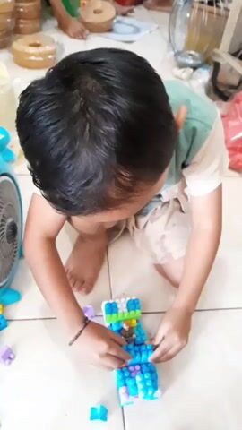 A child is kneeling on a tiled floor, focused on building with colorful blocks. The child wears a light green and white shirt and light-colored shorts. Their hands are occupied with assembling a structure made of blue, white, purple, and green blocks. Scattered around the child are individual blocks and a blue fan. In the background, out of focus, are stacked containers and a blurry figure. The lighting suggests an indoor setting.