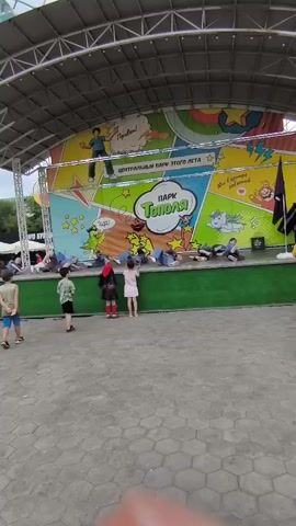 A group of children are performing a dance routine on a stage. They are dressed in matching blue outfits. The stage is decorated with colorful comic book-style graphics. A few adult audience members are watching the performance from the side. The event appears to be taking place outdoors, possibly in a park.