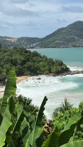 A scenic aerial view shows a lush green coastline with a crescent-shaped beach bordered by turquoise water and gentle waves. The beach has a few scattered rocks and some people are visible on the sand. The shoreline is dotted with palm trees and dense vegetation, with buildings and more greenery lining the hills in the background. The sky is partly cloudy. Banana leaves frame the foreground of the shot.