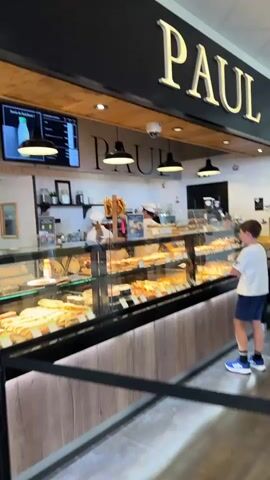 The video shows a bakery with pastries and sandwiches on display. The sign above the counter reads "PAUL." There are cameras on the ceiling. A person in a white shirt is working behind the counter. There are several customers in the store, some queuing and others looking at the merchandise. The environment appears to be a modern retail space with clean white walls and wooden accents.