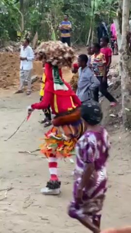 The video shows a person in costume dancing for a crowd of people outside, possibly in a village. The dancer wears red clothing with white and red striped leggings, and a colorful fringed skirt. They have a large, brown headdress with a green and white scarf. The dancer carries a small red object in their hand. The crowd consists of both children and adults, dressed in various casual clothes. The environment appears to be a dirt path with lush green vegetation and trees in the background. There is a dirt pile in the background as well.