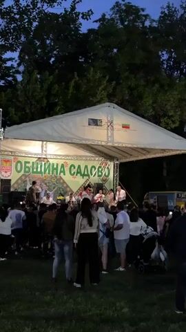 The video shows a gathering of people in an outdoor park area at night. A stage with musicians performing is visible, along with a sign in Bulgarian. The crowd is gathered in front of the stage. In the background, there are illuminated food stalls and a carousel. A man in a dark jacket is seen dancing alone, while another man is standing nearby. The overall atmosphere suggests a festival or public event taking place at night.