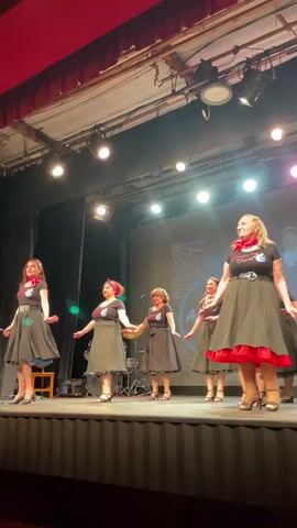 The video shows six women on a stage performing a dance routine. They are all wearing similar outfits consisting of black dresses with red trim and scarves around their necks. The stage has a backdrop with a stylized design and some lighting. Some of the women have their arms extended, while others are clapping. There is a drum set visible behind one of the dancers, suggesting a live band may be present off-camera. The lighting on stage is a mix of white and pink lights. The overall atmosphere appears to be a live performance in a theater or auditorium.