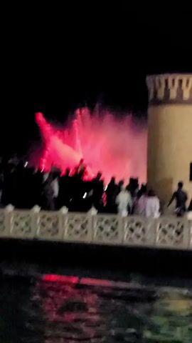 This media file captures a nighttime outdoor event featuring a water fountain show. A crowd of people, silhouetted against the illuminated fountain, stands behind a decorative white railing. The fountain itself is the main focus, erupting with plumes of water that are vividly colored with changing lights, shifting from pink to purple to various shades of blue.

To the right of the frame, a tall, cylindrical tower with a patterned top stands prominently. Its textured surface is lit, suggesting it is a man-made structure, possibly part of a landmark or attraction. The overall scene is dark, indicating it is nighttime, and the focus on artificial lighting for the fountain and the tower suggests an orchestrated event designed for spectacle.

The presence of the crowd gathered to watch the fountain implies a communal experience, likely one of enjoyment and wonder as they observe the dynamic water display. The emotions conveyed are primarily positive, centered around entertainment and shared visual experience. There are no visible text elements in the media file. The context provided places this event in Satkak, Uzbekistan, which might suggest a cultural or recreational gathering specific to that region.