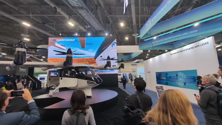 The video shows a crowded indoor convention hall.  In the foreground, there is a white and black electric vertical takeoff and landing (eVTOL) aircraft on display on a dark platform. The door of the aircraft is open, and there are people standing around looking at it and taking photos.  Behind the aircraft is a large screen showing the aircraft in flight.  The booth has a sign that says "SAIRO MOTORS GROUP."  Further back, on the right, is another booth displaying information about LiDAR technology. The environment appears to be a trade show or exposition, likely focused on technology and transportation. There are people walking around and interacting with the exhibits. The ceiling of the hall is high, with exposed industrial infrastructure. Banners hanging from the ceiling read "SOS Smart Optical Sensor LiDAR" and "We make the World Smarter with LiDAR."