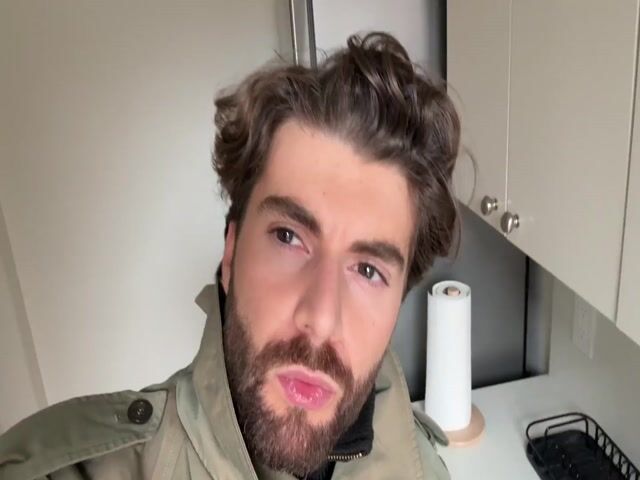 A man with brown hair and a beard is looking directly at the camera. He is wearing a green jacket and a black turtleneck. He tilts his head slightly and smiles. The background includes a white wall and kitchen cabinets with chrome hardware. A chrome ventilation hood is also visible. A small black drying rack sits on a countertop.