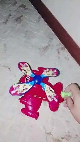 A hand is seen pushing a green lever on a red toy airplane that has a propeller. The propeller begins to spin rapidly, causing the toy to move across a patterned surface. The toy is bright red with a blue center on the propeller and has colorful designs on the propeller blades.