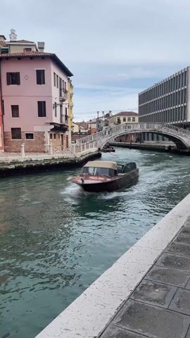 A boat is moving along a canal in Venice, Italy. The canal is surrounded by buildings of varying heights and architectural styles. There is a white, arched bridge in the background, and another boat passes underneath the bridge. The water is green, and the sky is overcast. The boat in the foreground is brown with a beige canopy. The perspective is from the side of the canal. The camera is still.