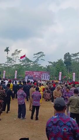 The video shows a large outdoor gathering with a crowd of people. In the background, there is a stage with a banner that reads "DIRGAHAYU INDONESIA" and features a group photo of people. Several Indonesian flags are visible, including one flying on a tall pole and smaller ones on poles along the edge of the gathering. The environment appears to be a grassy or dirt field surrounded by trees. People are dressed in various outfits, some in uniforms, others in casual clothing or traditional attire. The crowd is looking towards the stage, and some individuals are taking photos or videos with their phones. The sky is partly cloudy.
